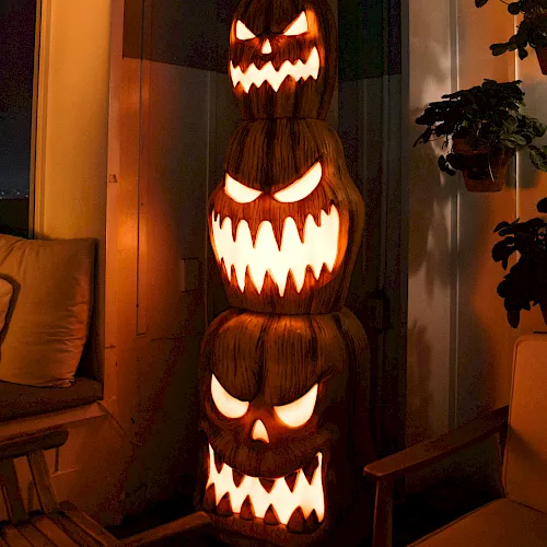 A glowing carved jack-o&rsquo;-lantern monster with sharp teeth sits indoors beside a couch and potted plants, lighting the room up.