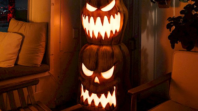 A glowing carved jack-o&rsquo;-lantern monster with sharp teeth sits indoors beside a couch and potted plants, lighting the room up.
