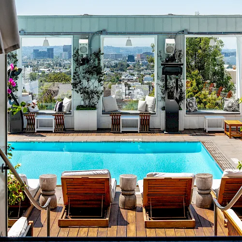 An urban rooftop patio with a bright blue pool, lounge chairs, potted plants, and a city skyline view beyond tall windows.