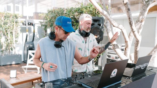Two DJs wearing headphones and a cap work a DJ mixer at an outdoor event, while a man with his arms out monitors audio.