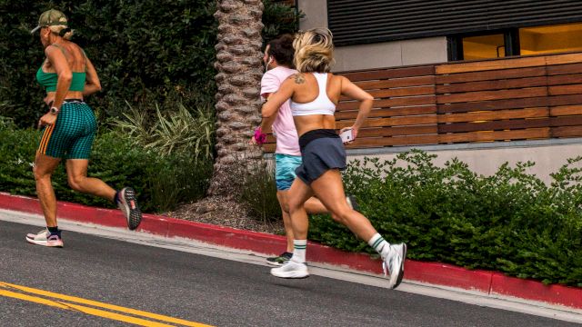 Three runners sprint on an outdoor track beside a stone pillar and greenery.