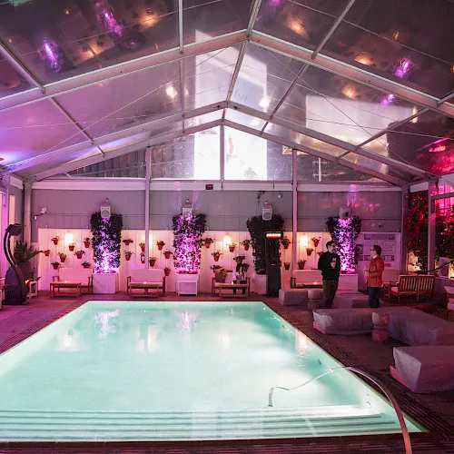 An indoor pool event under a glass roof, glowing purple lights, potted plants, and a couple standing nearby.